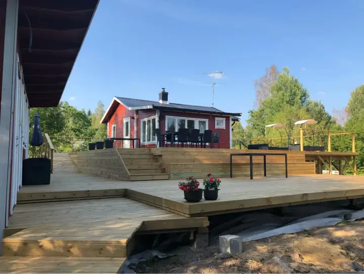 Stor altan i trä med trappor och sittplatser framför ett rött hus. Perfekt altan inspiration för sommarens grillkvällar.