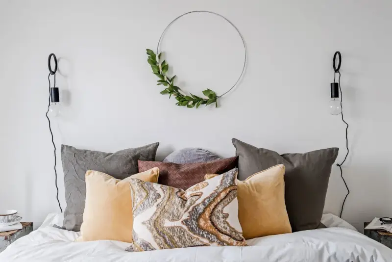 Sovrum inspiration med en säng fylld av kuddar i olika färger och texturer, en grön krans på väggen och minimalistiska vägglampor.
