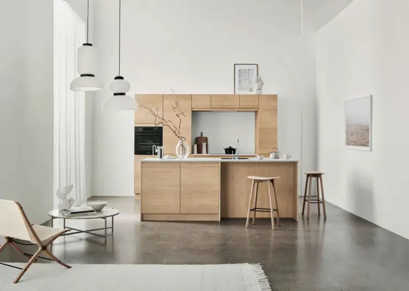 Ett modernt kök med minimalistisk design i ljust trä. Tre papperslampor hänger från taket. En fåtölj och ett runt soffbord står till vänster.